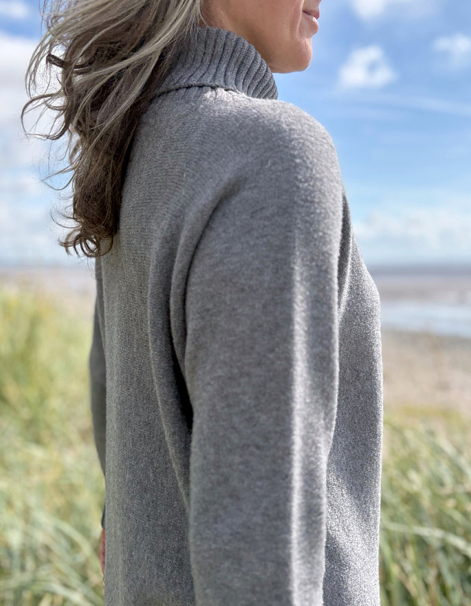 Person wearing a grey turtle neck sweater and sunglasses standing in a grassy field with a blue sky.