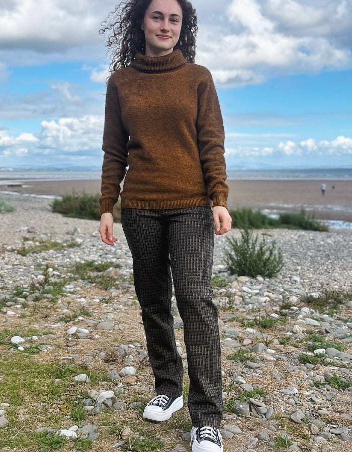 A person standing outdoors on a rocky, grassy shoreline wearing dark patterned plaid check trousers, a brown knit sweater, and black-and-white sneakers.