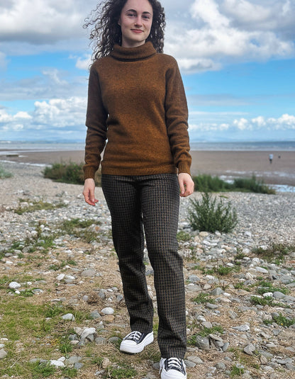 A person standing outdoors on a rocky, grassy shoreline wearing dark patterned plaid check trousers, a brown knit sweater, and black-and-white sneakers.