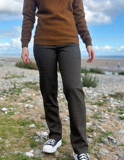 A person standing outdoors on a rocky, grassy shoreline wearing dark patterned plaid check trousers, a brown knit sweater, and black-and-white sneakers.
