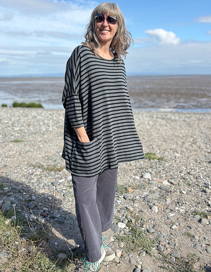 Woman standing on a pebbly beach wearing a striped black and grey oversized sweatshirt top  and sunglasses.