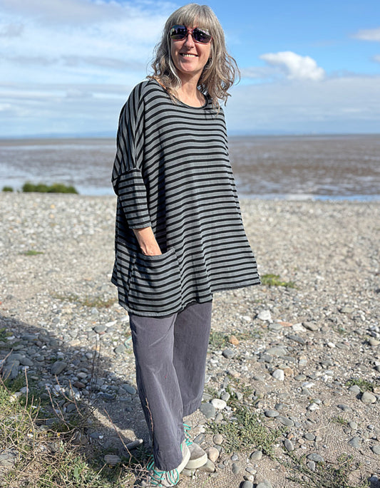 Woman standing on a pebbly beach wearing a striped black and grey oversized sweatshirt top  and sunglasses.