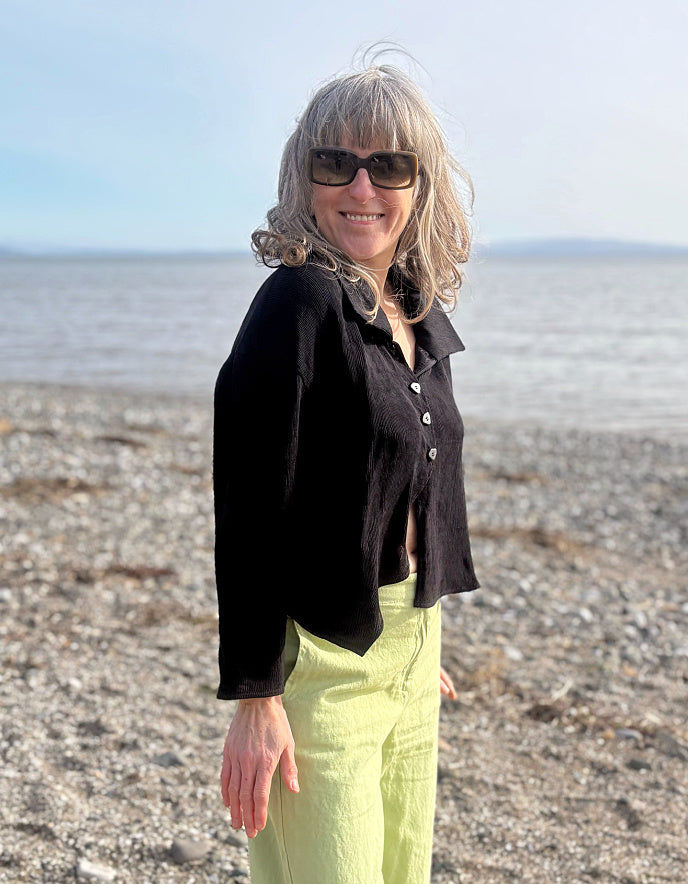 Woman standing on a beach wearing sunglasses and a black jacket.