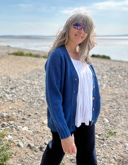 Woman wearing a wool blue cardigan on a pebbly beach with water and sky in the background