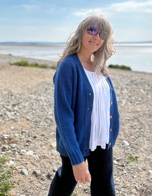 Woman wearing a wool blue cardigan on a pebbly beach with water and sky in the background