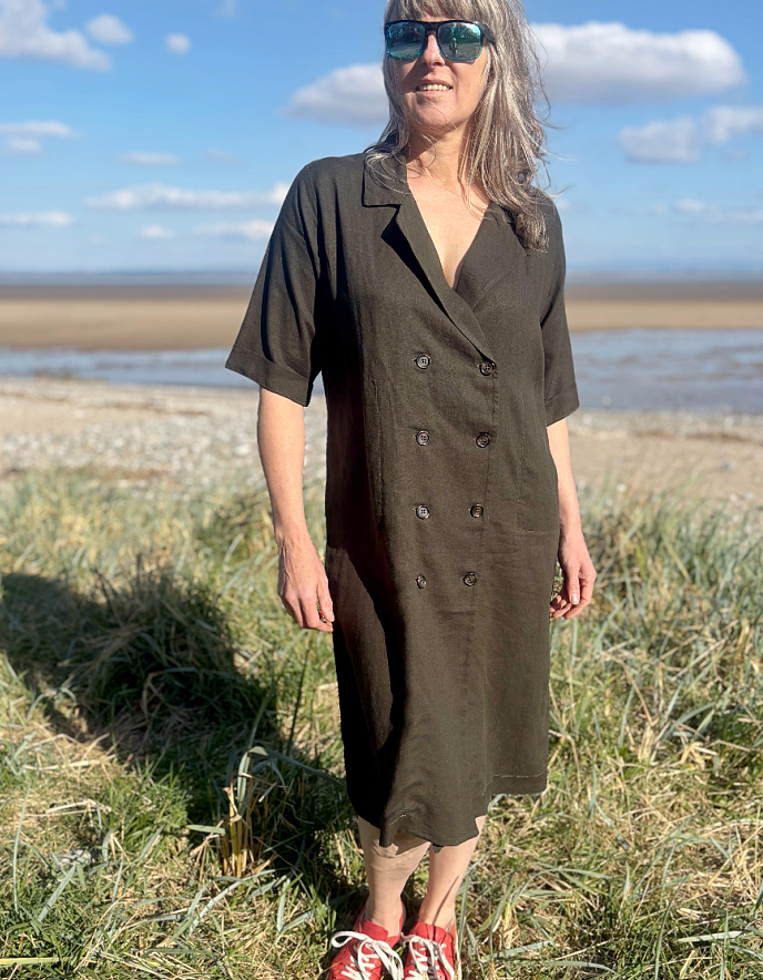 Woman in a long dark green shirt dress standing on a grassy field with a coastal background.