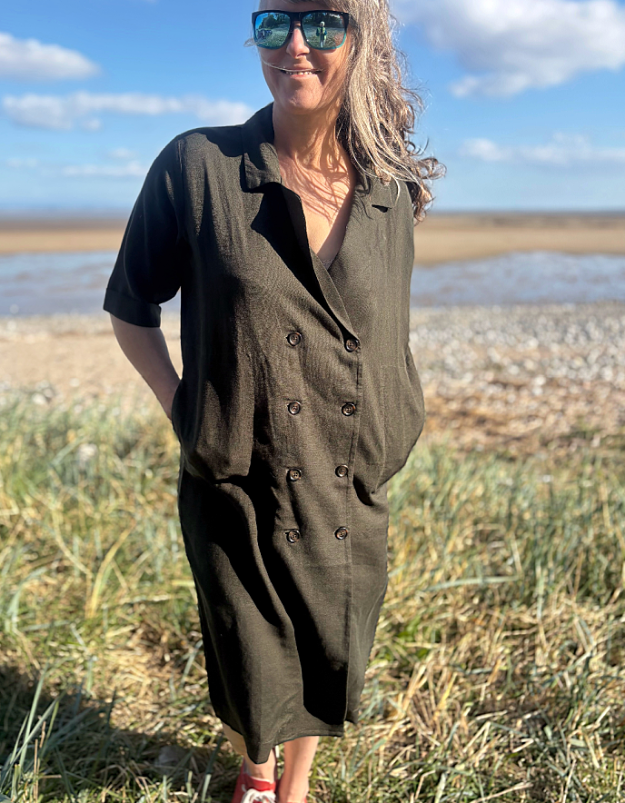 Woman in a long dark green shirt dress standing on a grassy field with a coastal background.