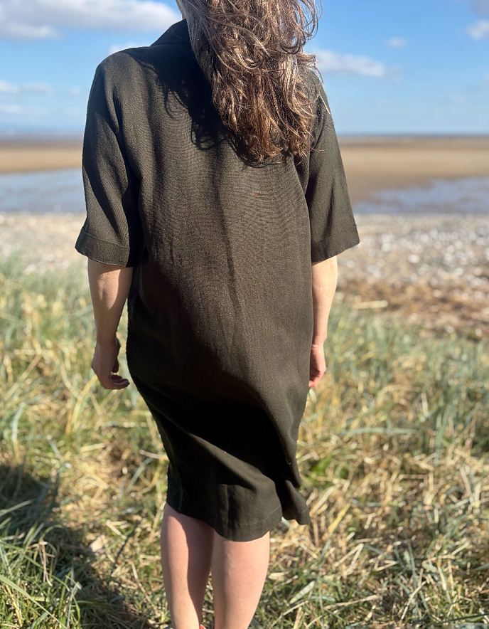 Woman in a long dark green shirt dress standing on a grassy field with a coastal background.