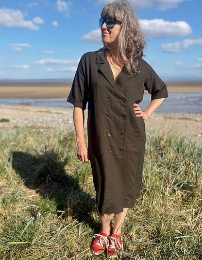 Woman in a long dark green shirt dress standing on a grassy field with a coastal background.