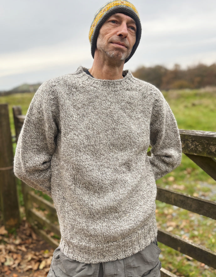 Man wearing a gray knitted sweater and yellow beanie standing outdoors with a wooden fence and grassy field in the background.