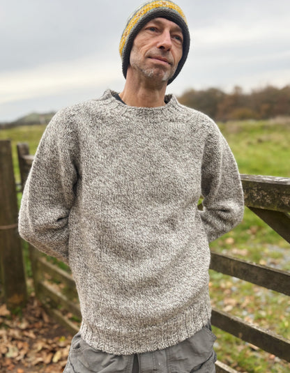 Man wearing a gray knitted sweater and yellow beanie standing outdoors with a wooden fence and grassy field in the background.