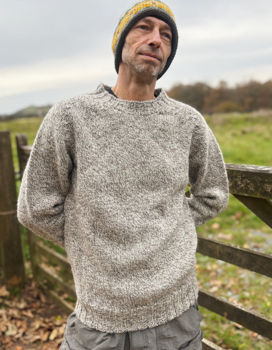 Man wearing a gray knitted sweater and yellow beanie standing outdoors with a wooden fence and grassy field in the background.