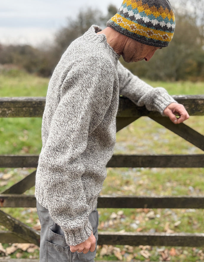 Person wearing a patterned knit sweater and hat leaning against a wooden gate outdoors.