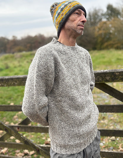 Man wearing a gray knit sweater and colorful beanie standing in a field with a wooden fence.