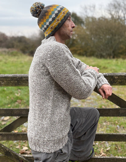 Person wearing a knitted sweater and hat sitting on a wooden fence outdoors.