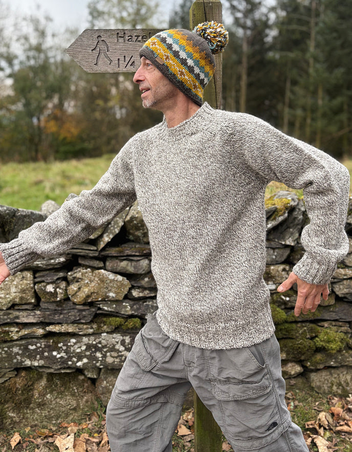 Man wearing a gray sweater and colorful knit hat standing next to a stone wall with a sign in the background.