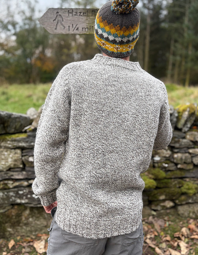 Person wearing a gray knitted sweater and colorful beanie standing in a natural setting with a stone wall and sign in the background.
