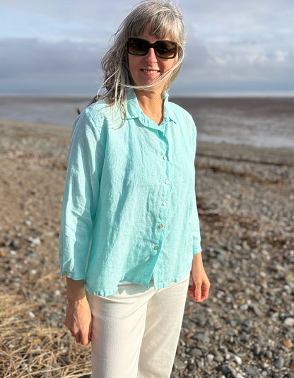 Woman in a light blue shirt and sunglasses standing on a pebbly beach with ocean in the background