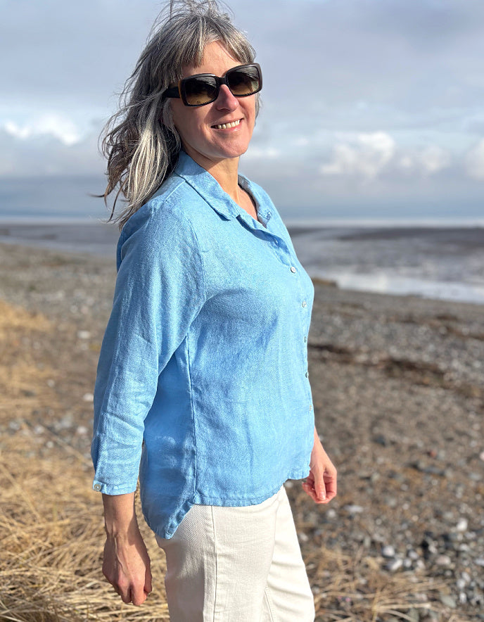 Woman wearing a light blue shirt and sunglasses standing on a beach with ocean and sky in the background.