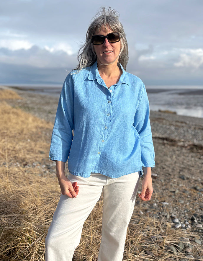 Woman wearing a light blue shirt and sunglasses standing on a beach with ocean and sky in the background.