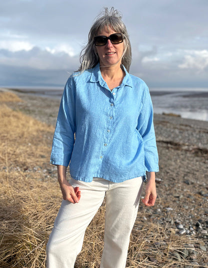 Woman wearing a light blue shirt and sunglasses standing on a beach with ocean and sky in the background.