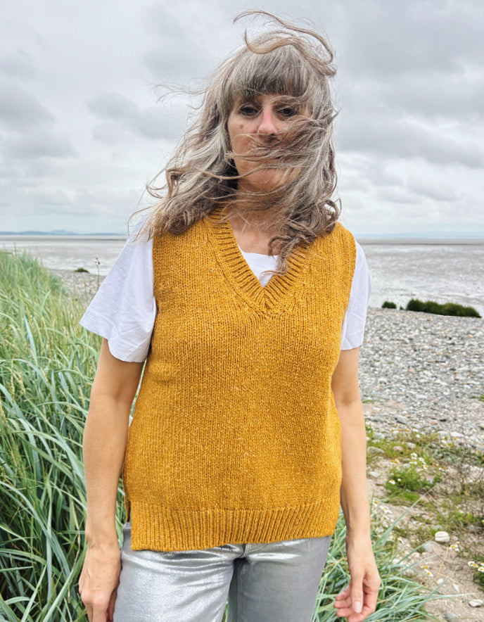 Person wearing a mustard yellow knitted vest over a white shirt, standing outdoors with grass and water in the background.