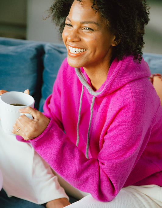Woman in a pink hoodie holding a mug, sitting on a couch.