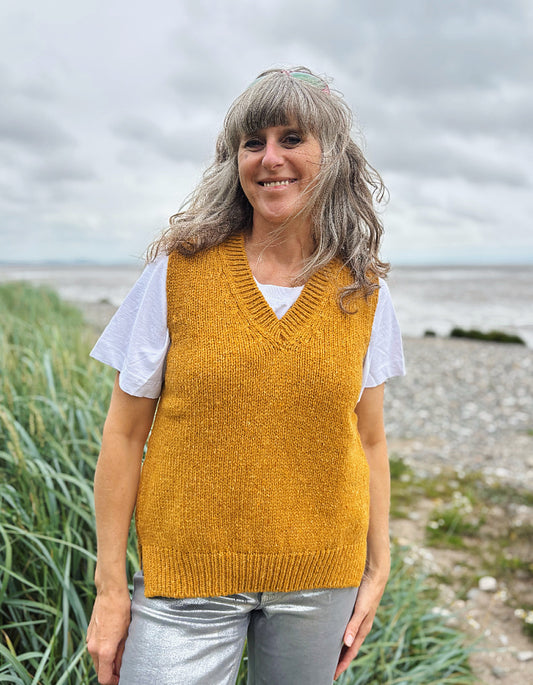 Woman wearing a mustard yellow knitted vest over a white shirt, standing outdoors with a natural background.