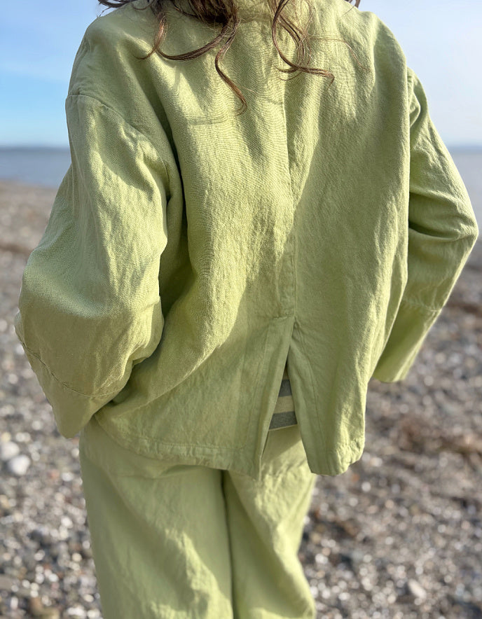 Person wearing a light green cotton linen pant suit standing on a pebbly beach.