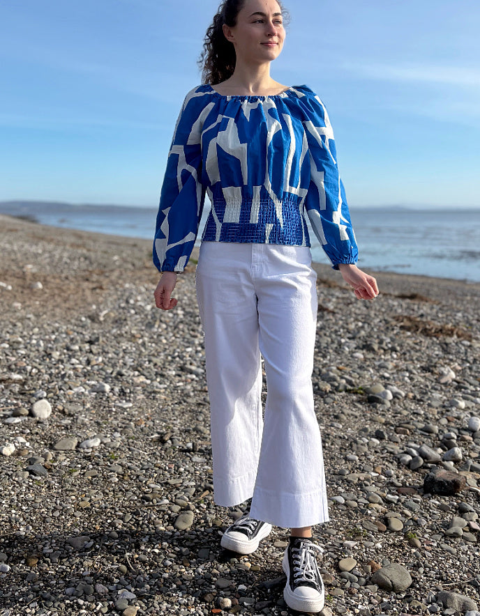 Woman wearing a blue and white patterned top and white pants standing on a pebbly beach.