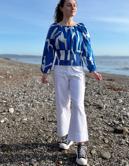 Woman wearing a blue and white patterned top and white pants standing on a pebbly beach.