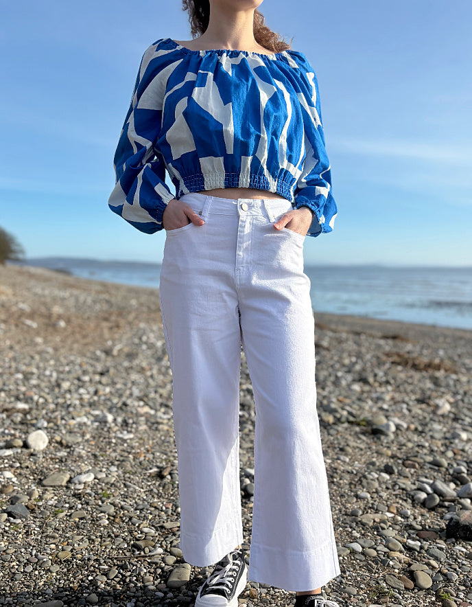 Person wearing a blue and white patterned top and white pants standing on a pebbly beach.