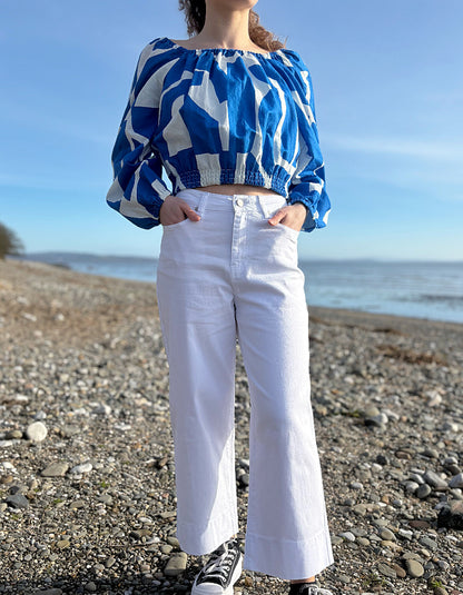 Person wearing a blue and white patterned top and white pants standing on a pebbly beach.