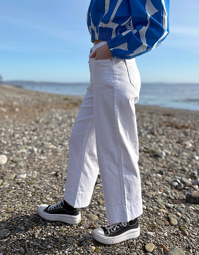 Person wearing a blue patterned shirt, white pants, and black sneakers on a pebbly beach.