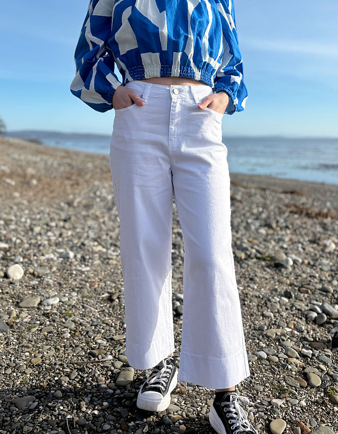 Person wearing a blue and white patterned shirt, white pants, and black sneakers on a pebbly beach.
