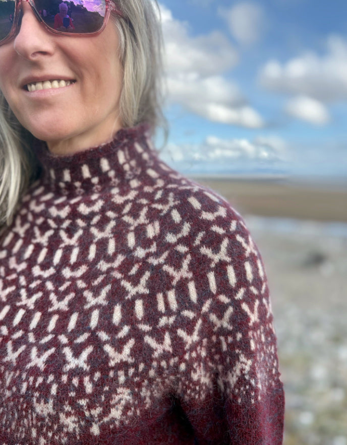 Person wearing a Icelandic burgundy patterned sweater standing on a beach with a scenic background