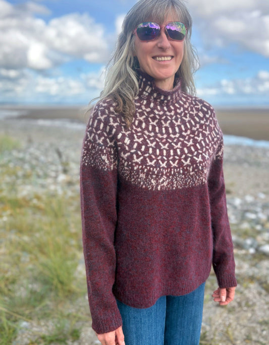 Person wearing a Icelandic burgundy patterned sweater standing on a beach with a scenic background