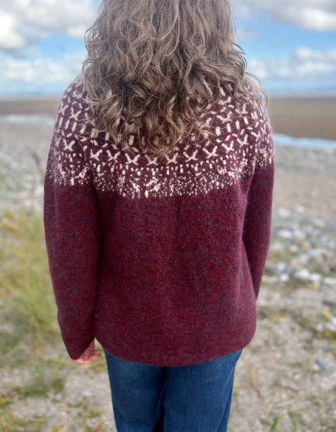 Person wearing a Icelandic burgundy patterned sweater standing on a beach with a scenic background