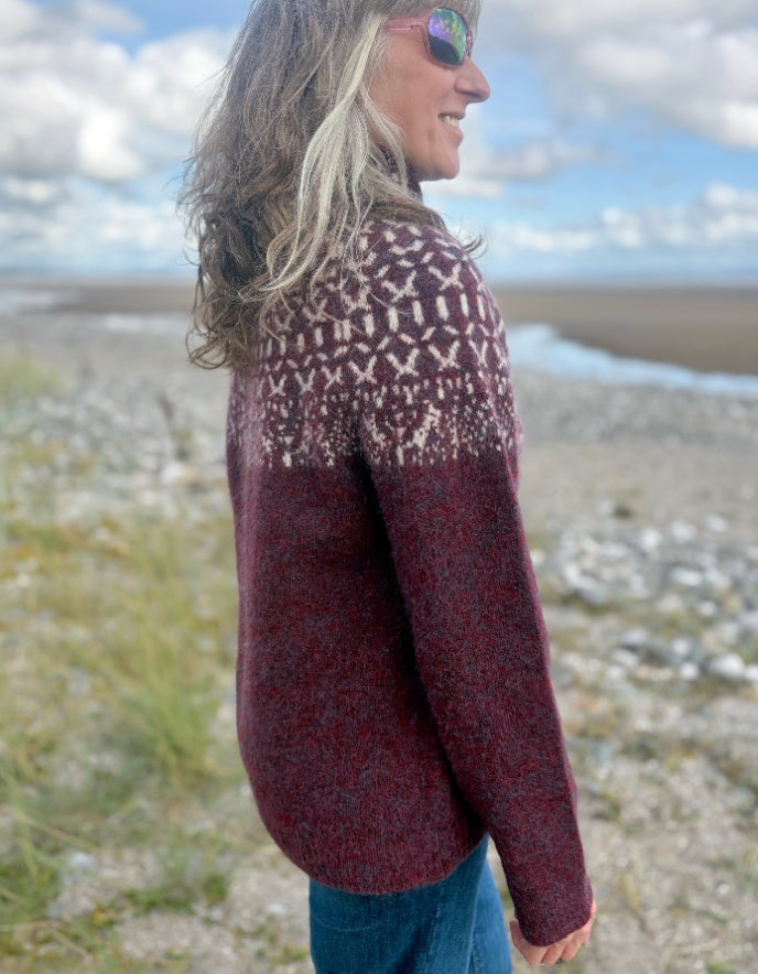 Person wearing a Icelandic burgundy patterned sweater standing on a beach with a scenic background