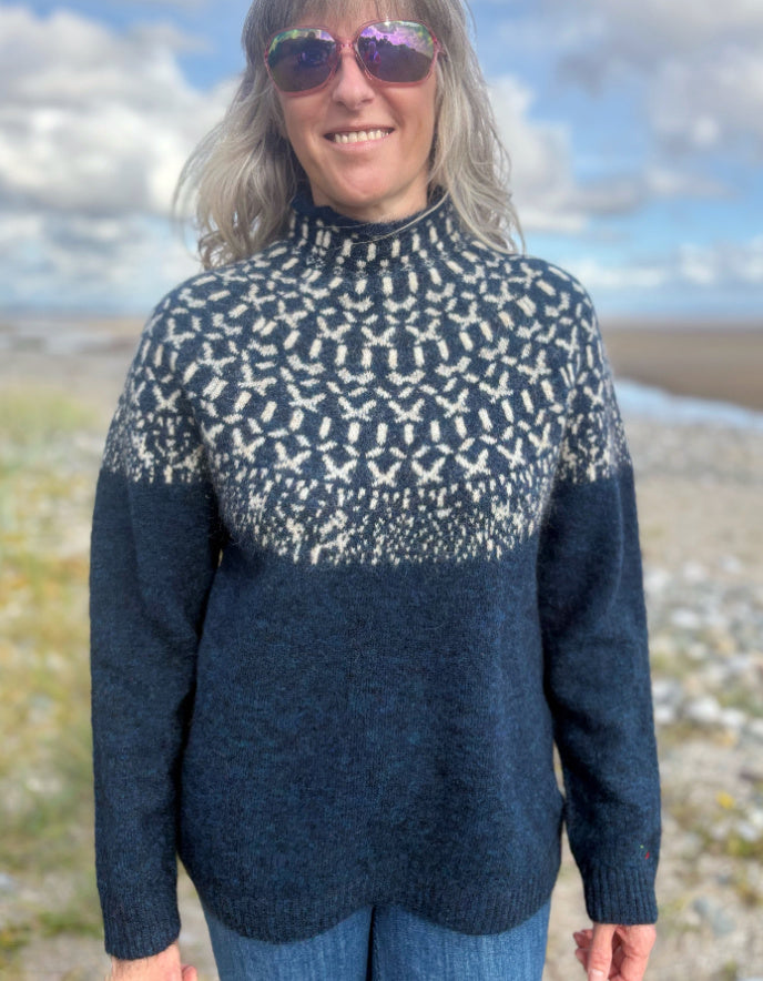 Person wearing a patterned navy sweater on a beach with a blue sky.
