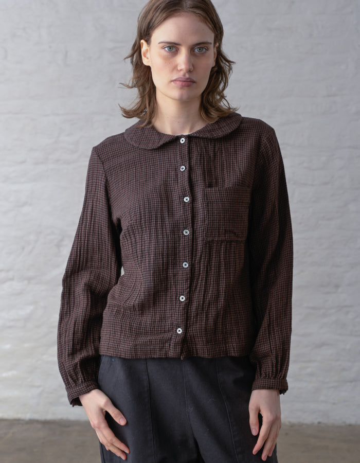 A woman wearing a dark brown and black textured gingham long-sleeve button-up shirt with a Peter Pan collar, standing against a light gray brick wall.