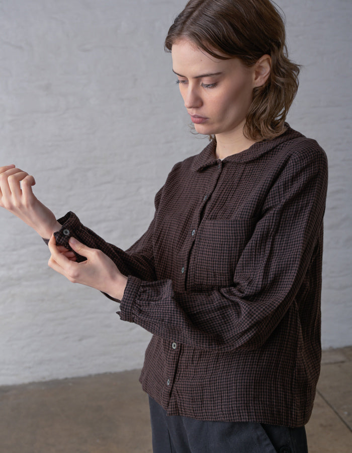 A woman wearing a dark brown and black checkered long-sleeve shirt, adjusting the cuff of the sleeve against a light gray brick wall background.