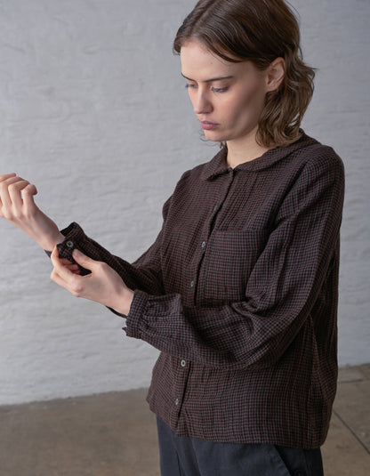 A woman wearing a dark brown and black checkered long-sleeve shirt, adjusting the cuff of the sleeve against a light gray brick wall background.