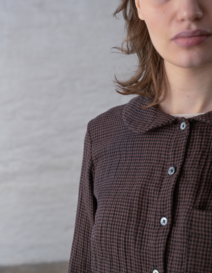 Close-up of a person wearing a brown and black checkered button-up shirt with a Peter Pan collar, photographed against a light textured wall.