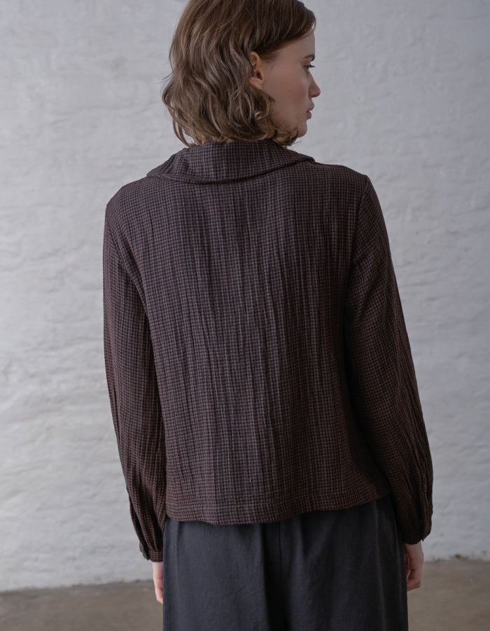 A woman shown from the back wearing a dark brown and black textured gingham long-sleeve jacket with a high folded collar, paired with dark gray trousers, set against a white textured wall.