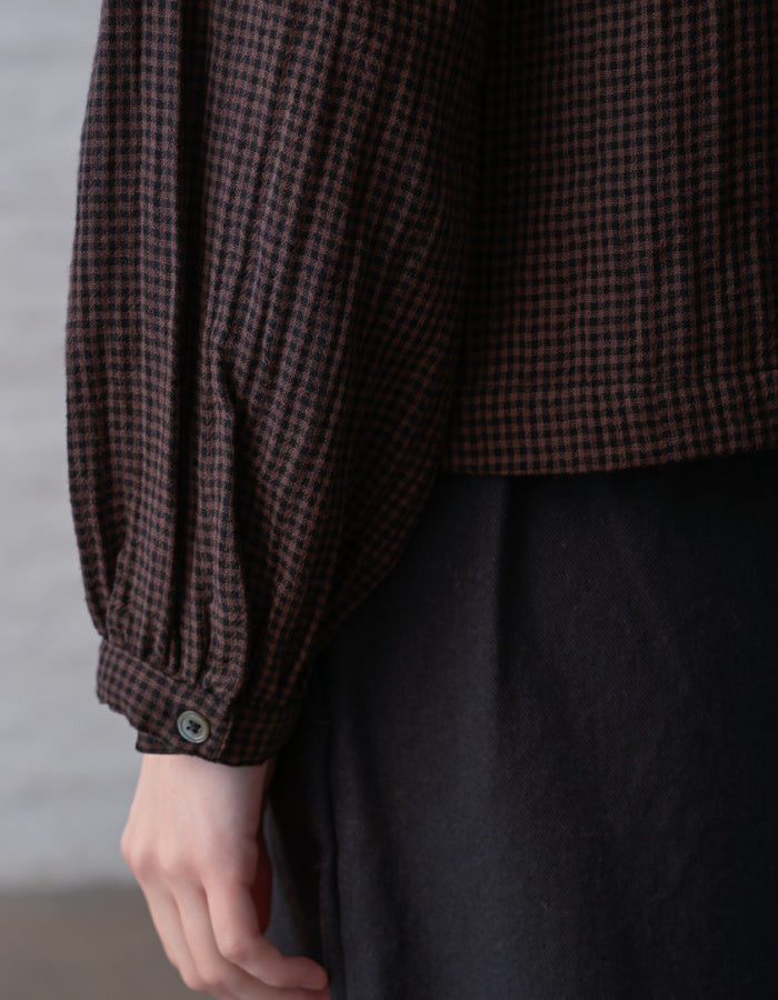 Close-up of a person wearing a dark brown and black checkered long-sleeve blouse with gathered cuffs and a dark skirt.