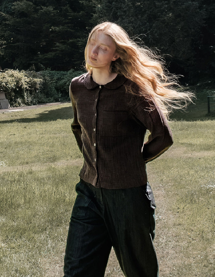 A woman standing outdoors in a grassy park area, wearing a dark brown textured button-up shirt with a collar and dark wide-leg pants.