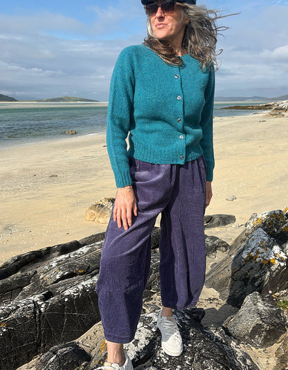 A woman standing on rocks by a beach wearing a teal button-front cardigan, purple corduroy pants, and white sneakers.