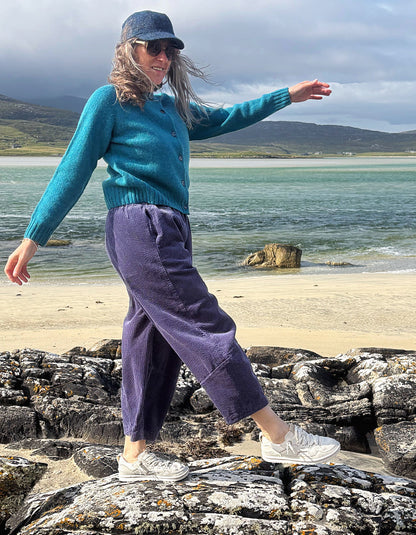 A woman stands on rocks by the beach wearing a teal knit cardigan, purple corduroy pants, a blue bucket hat, sunglasses, and white sneakers, with the sea and hills in the background.