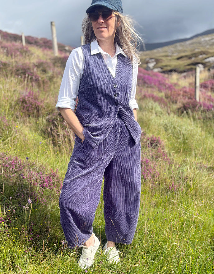 A woman standing outdoors in a grassy field with purple heather and hills in the background, wearing a dark blue corduroy vest and matching wide-leg trousers over a white collared shirt, with a dark cap and white sneakers.